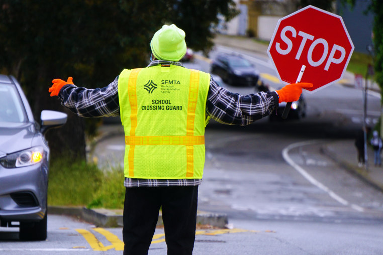The back of a crossing guard with a yellow vest and stop sign raises their arms to signal to an oncoming car.