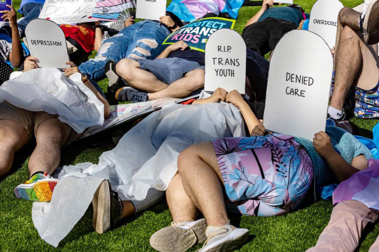 Protesters lie on the ground holding cardboard signs shaped like tombstones that read "Depression," "R.I.P. Trans Youth," "Denied Care" and "Overdosed"