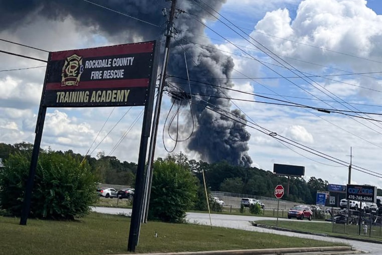 Dark smoke rises into the sky from behind trees