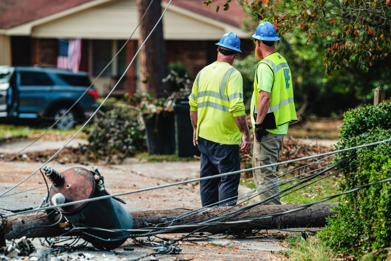 Two workers wearing hardhats survey damage on a street near a fallen electrical pole