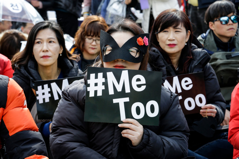 A person wears a black mask and holds a sign that says "#Me Too" in a crowd of protesters