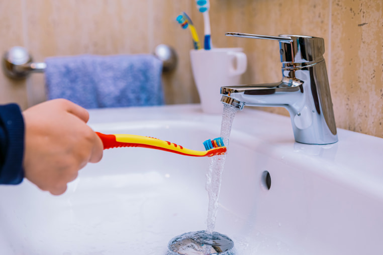 Detail of the arm of a girl in the bathroom, pouring water on her toothbrush in the sink.