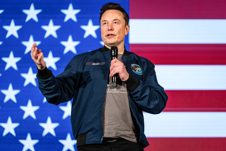 Elon Musk speaks on stage, with an American flag backdrop behind him