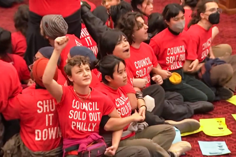 Protestors sit on the ground and link arms wearing shirts that say "Council Sold Out Philly"