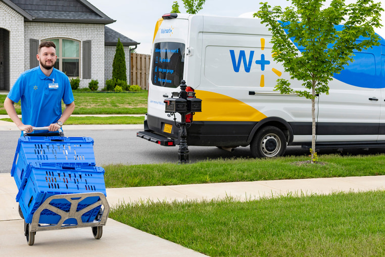 A man rolls two blue bins of items on a sidewalk near a parked Walmart delivery van