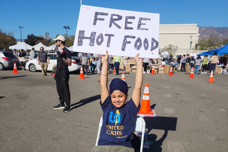 A boy sits in a chair and holds up a sign that says "Free Hot Food" outside