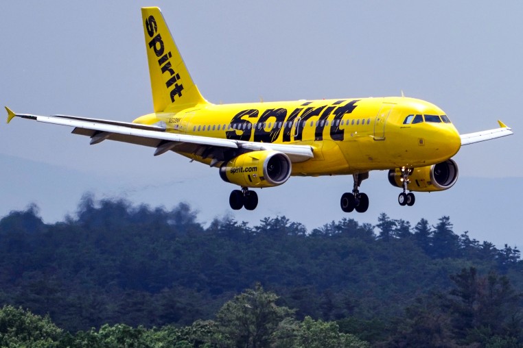 A yellow Spirit Airlines plane flies in the sky above trees