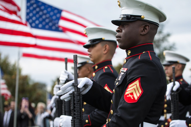 Marines stand outside in uniform and hold rifles, American flag flies in the background