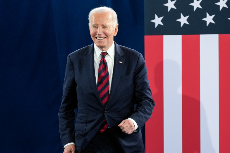 Joe Biden smiles and walks on stage, an American flag backdrop behind him