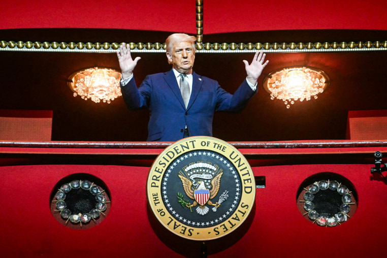 Donald Trump, with both hands raised, stands on a balcony inside of a building