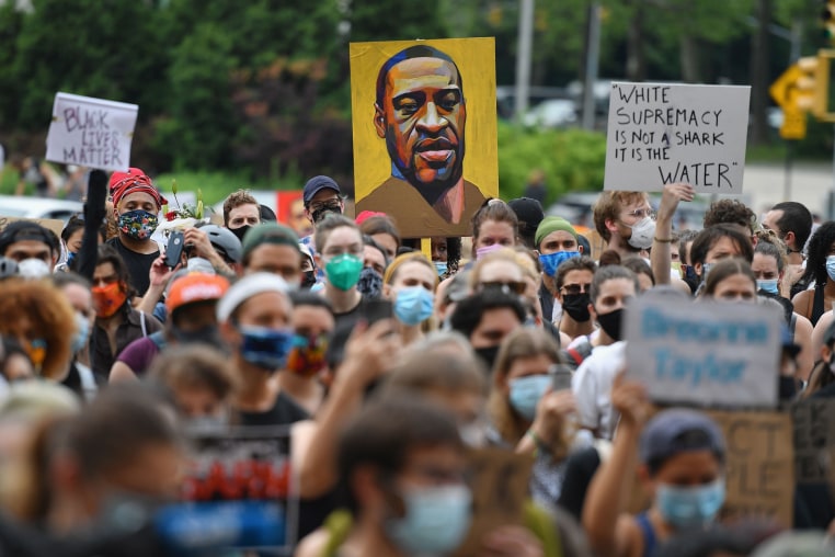 A big crowd at an outdoor protest, in the back is a large poster of George Floyd