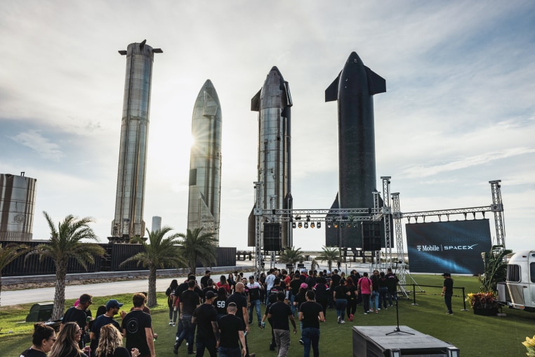 A crowd stands on a lawn in front of a stage, four large spaceship prototype sculptures in the background