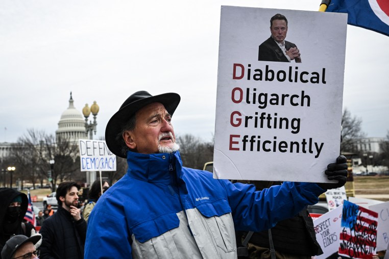A person holds a sign outside that reads "Diabolical Oligarch Grifting Efficiently"