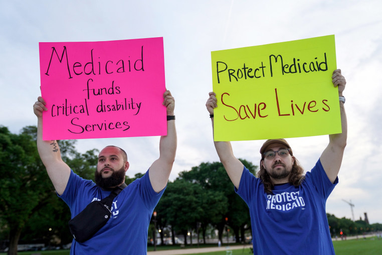 Two people hold signs outside, one the left it says "Medicaid funds critical disability services", and on the right it says "Protect Medicaid, Save lives"