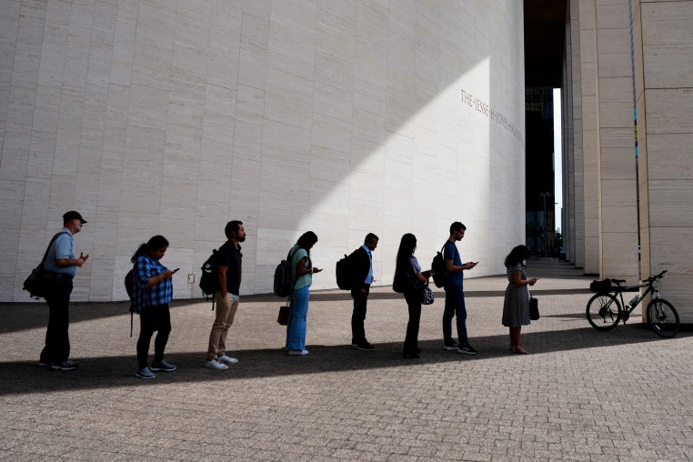 Eight people stand in a shadow to protect themselves from the sun.