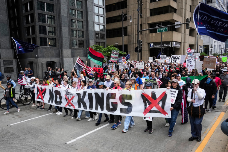 A large group of people walk through a street and hold a banner that says "No Kings"