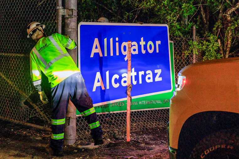 Two workers finish covering the Dade-Collier Training and Transition Airport sign with the "Alligator Alcatraz" sign