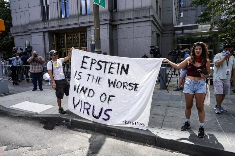 Two people hold a white blanket sign with black text that reads "Epstein is the worst kind of virus"