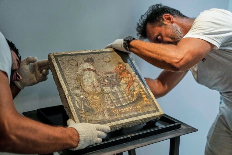 A mosaic artwork depicting a woman standing with her bum showing reaching out to a man who is laying on a bed underneath a blanket