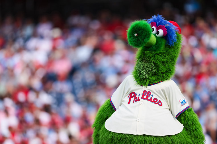 A fuzzy green mascot stands on the baseball field wearing a Phillies jersey