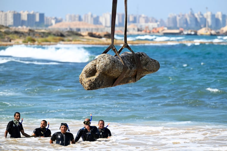 Divers watch as a crane pulls a piece of stone from the sea, with a cityscape in the background