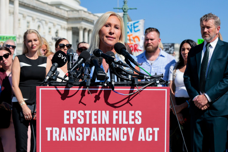 Marjorie Taylor Greene speaks at a podium with multiple microphones, people stand behind her, and the red sign in front of the podium reads "Epstein Files Transparency Act"