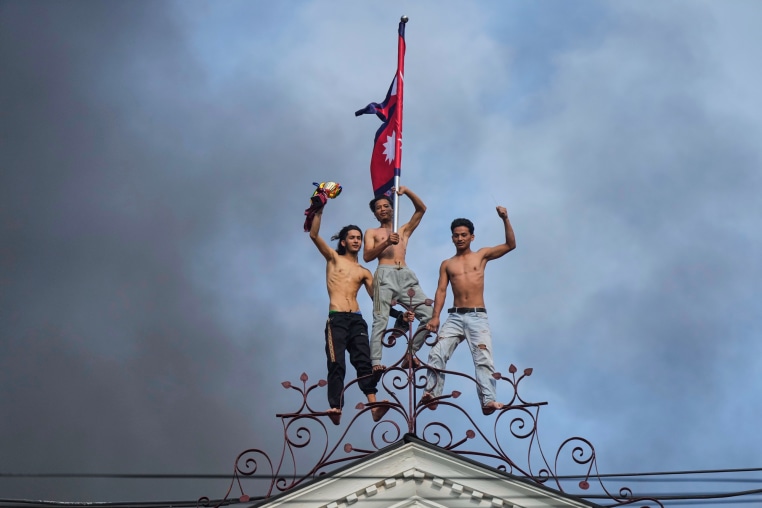 Three people stand on top of a building, the one in the center carries a Nepal flag