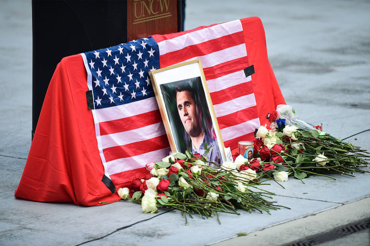 A framed photo of Charlie Kirk is laid against an American flag and surrounded by red and white roses on a memorial on the ground next to a podium