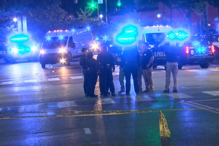 Police stand outside on the street in front of police cars with lights on, police tape is in front of them