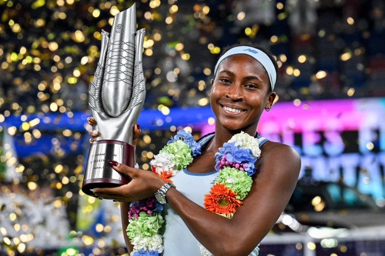 Coco Gauff holds a trophy while wearing a long flower necklace, gold confetti falls behind her