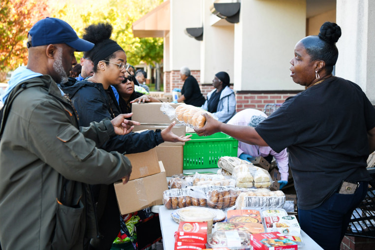 People in line at a food bank, a woman passes someone a bag of bagels from across a table lined with food