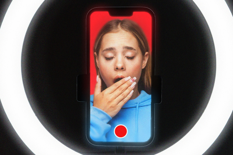 A girl yawning on a phone screen with a record button, surrounded by a ring light.