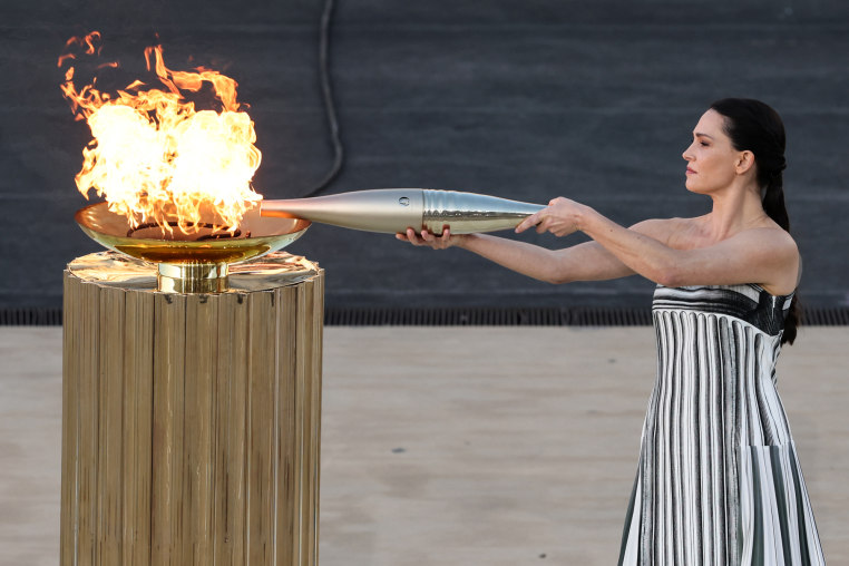 A woman lights a flame with large a silver torch in a golden dish atop a golden podium