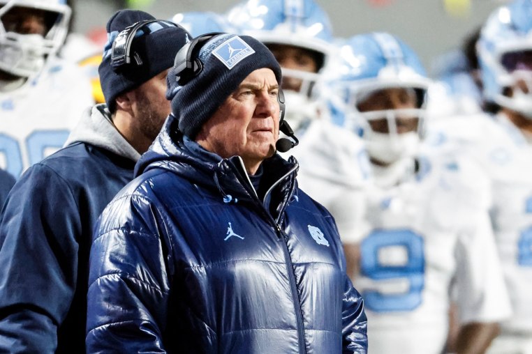 Bill Belichick stands on the sideline with players outside on the football field