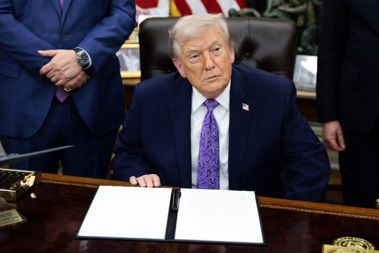 Donald Trump sits at his desk in the oval office, an executive order is on the desk