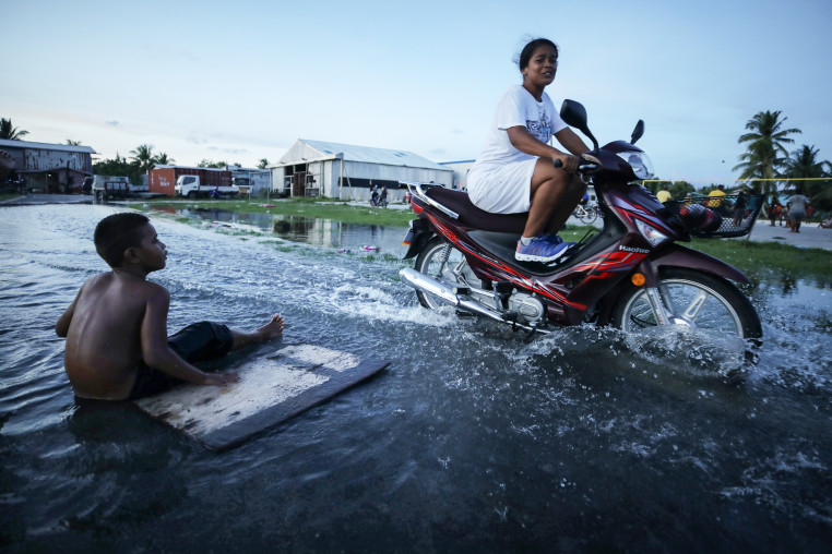 A woman rides her scooter through floodwaters next to a boy sitting near a plank on the flooded street