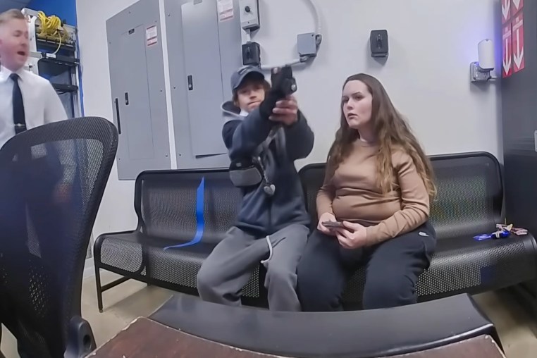 A man holds out a gun, attempting to shoot it, while seated on a couch next to a woman inside of a room. On the left, a guard yells and runs towards him