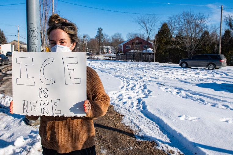 An activist holds a sign that reads "ICE IS HERE" near an alleged federal agent vehicle.