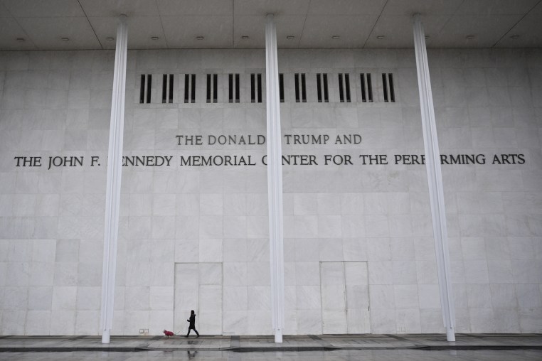 A person walks a dog in front of the tall facade of a building with lettering reading THE DONALD J. TRUMP AND THE JOHN F. KENNEDY MEMORIAL CENTER FOR THE PERFORMING ARTS.