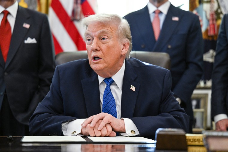 Donald Trump speaks while seated at his desk, hands clasped above an executive order on the desk