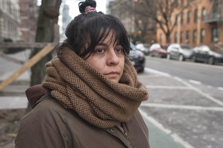 Val Calderon stands outside wearing a large chunky brown scarf and coat, a tear begins to stream down her face