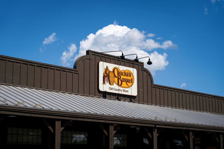 The orange and white Cracker Barrel logo is displayed on a brown building's facade, under clear blue skies.