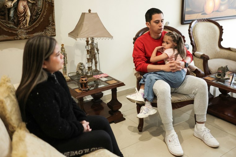 A father sits on a chair inside a living room with tiled flooring, holding his daughter in his lap.