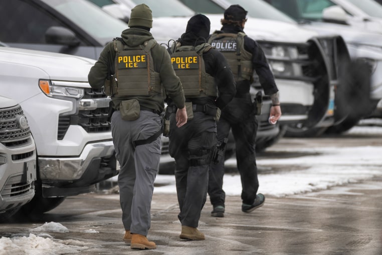 Three ICE agents wearing flak jackets walk over icy pavement with their backs turned to the camera.