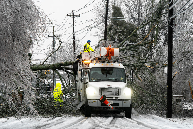 Two workers in blaze yellow suits work to clear fallen trees from a snow-covered roadway. 