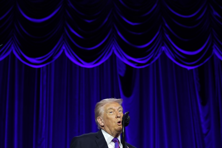 President Trump speaking into a microphone in front of a blue velvet stage backdrop.