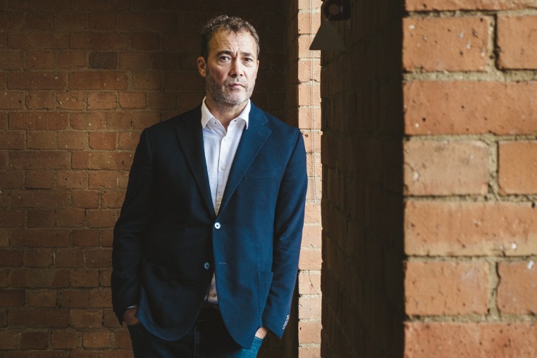 Will Lewis leans against a brick wall wearing a navy blazer and white shirt without a necktie.
