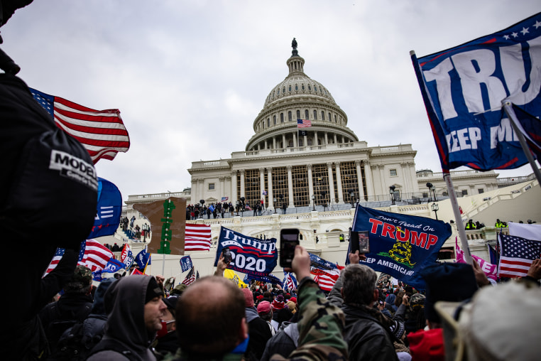 A huge crowd of people waving Trump 2020 flags are gathered outside the U.S. Capitol building under gray skies. 