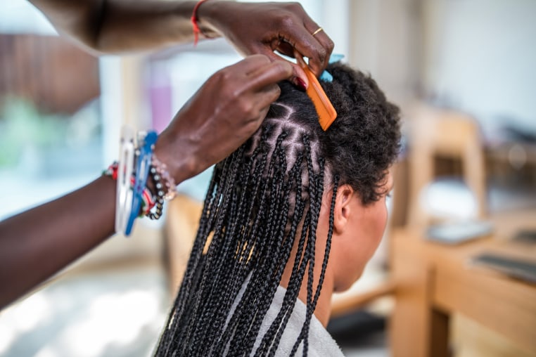 A mid adult Black woman braids the hair of a mixed race female in a domestic setting, showcasing a traditional hairstyle being made