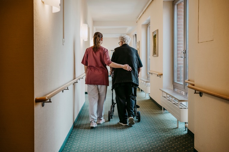 Rear view of female caregiver assisting senior woman walking with mobility walker in corridor